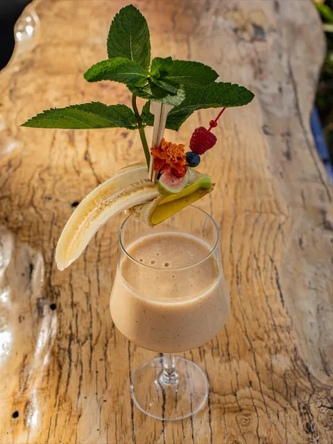 A banana cream cocktail topped with mint, marigold and a split banana, on a live-edge table.