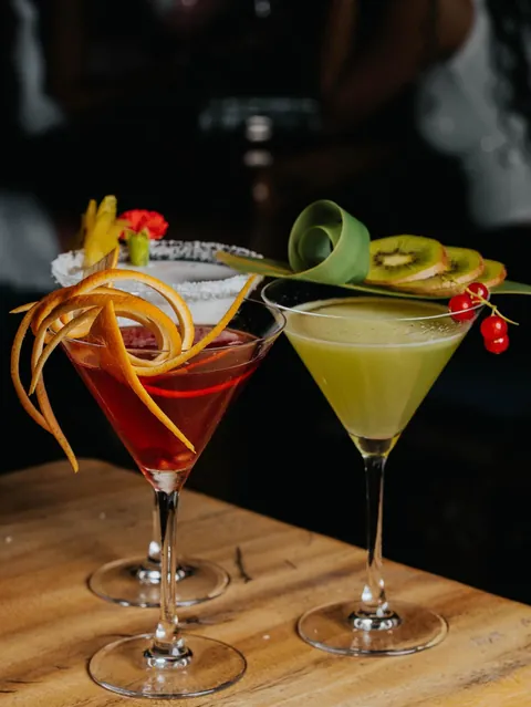 Two martinis side by side, one red with an orange peel spiral, one green with kiwi and redcurrants.