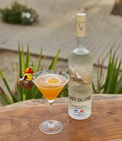A passion fruit martini garnished with edible flowers on a timber table at dusk.