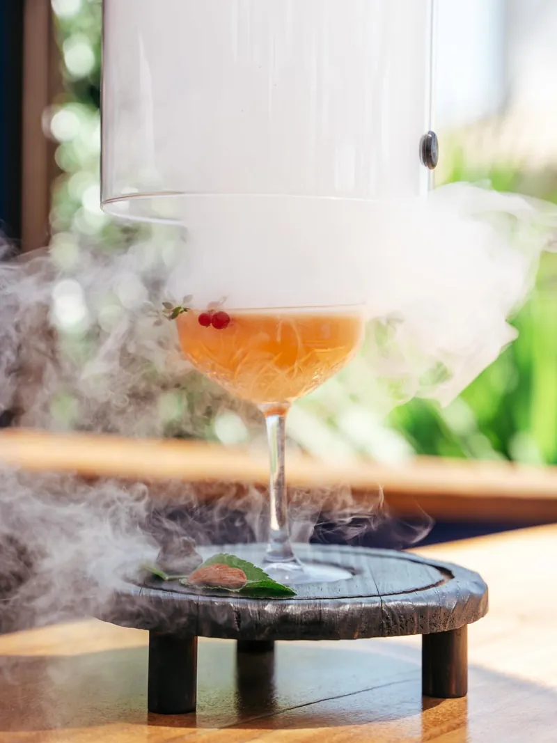 A glass cloche being lifted off a coupe cocktail, releasing a cloud of smoke across the table.