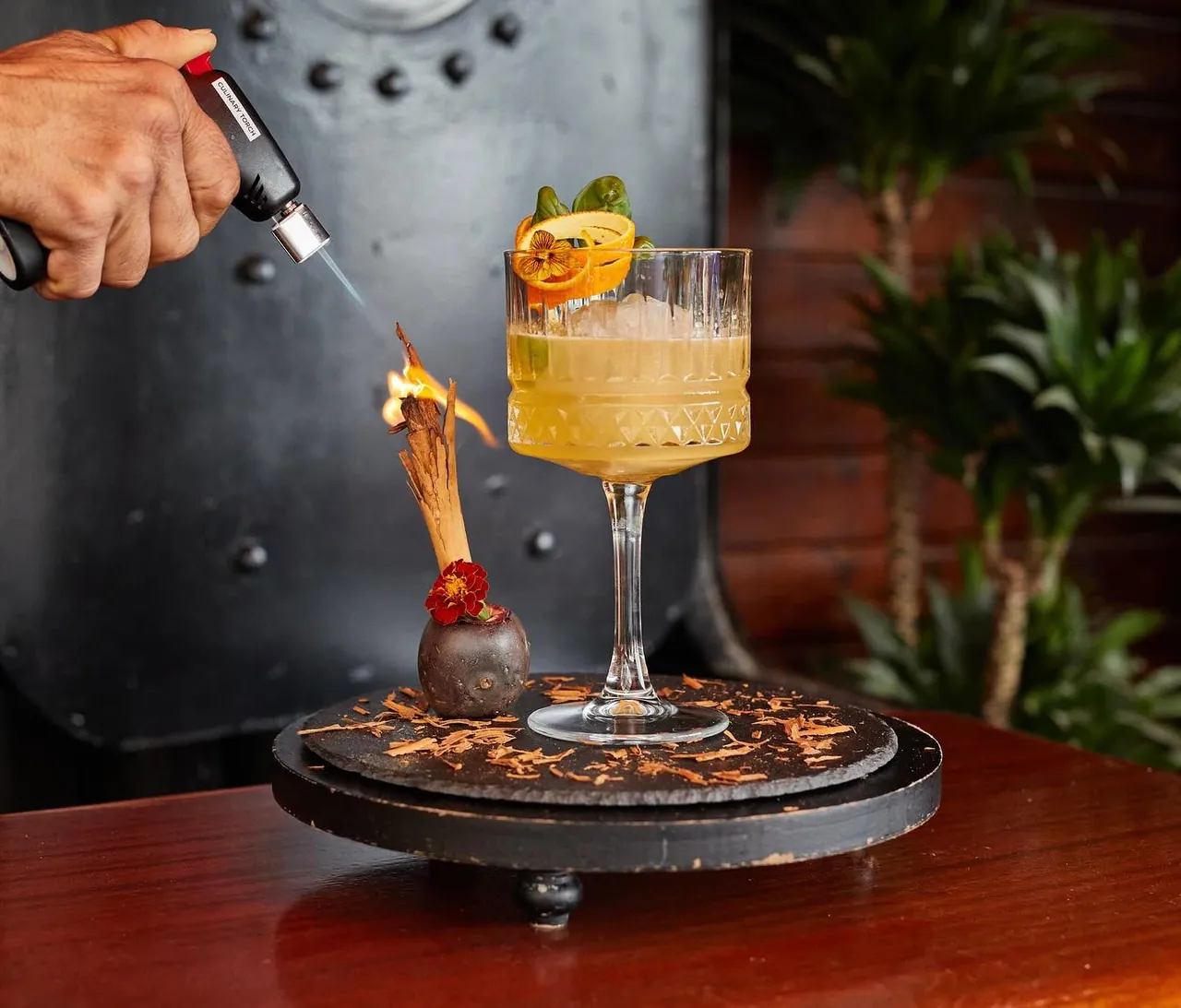 A bartender torching a cinnamon stick beside a finished cocktail on a black slate stand.