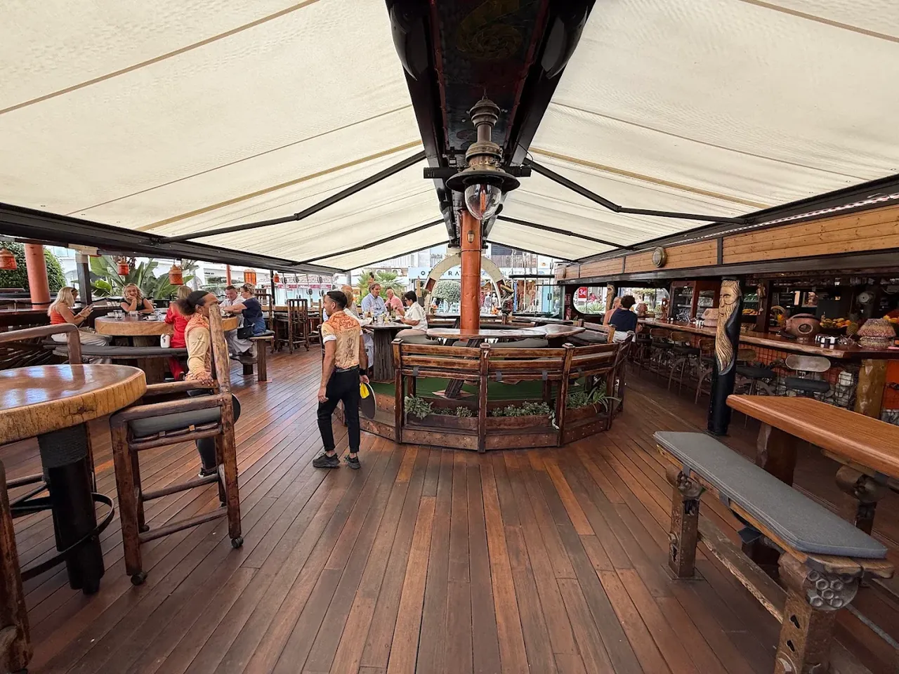 The main deck in daylight: broad timber planking, a mast through the centre, cream awnings overhead and guests at tables.