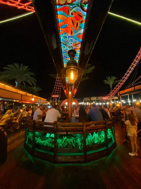 The deck at night, packed with people, lit by green uplights, an amber lantern and red-lit rigging.