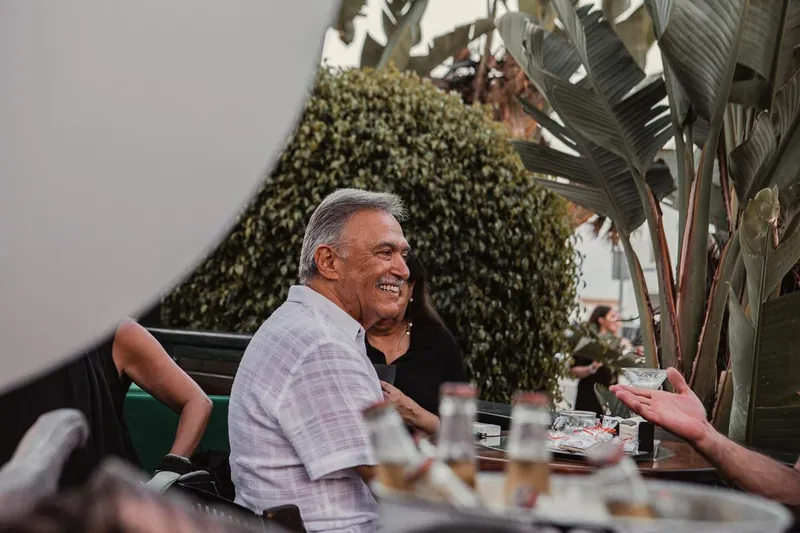A guest laughing at a table on the deck, with strelitzia and clipped hedges behind.