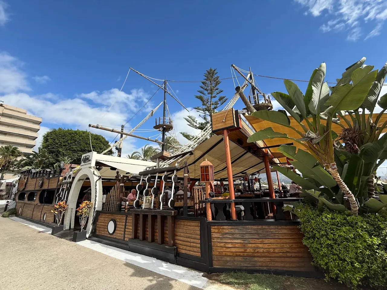 The Astral galleon seen from the street in Puerto Banús: a timber hull with masts, rigging and cream awnings against a blue sky.
